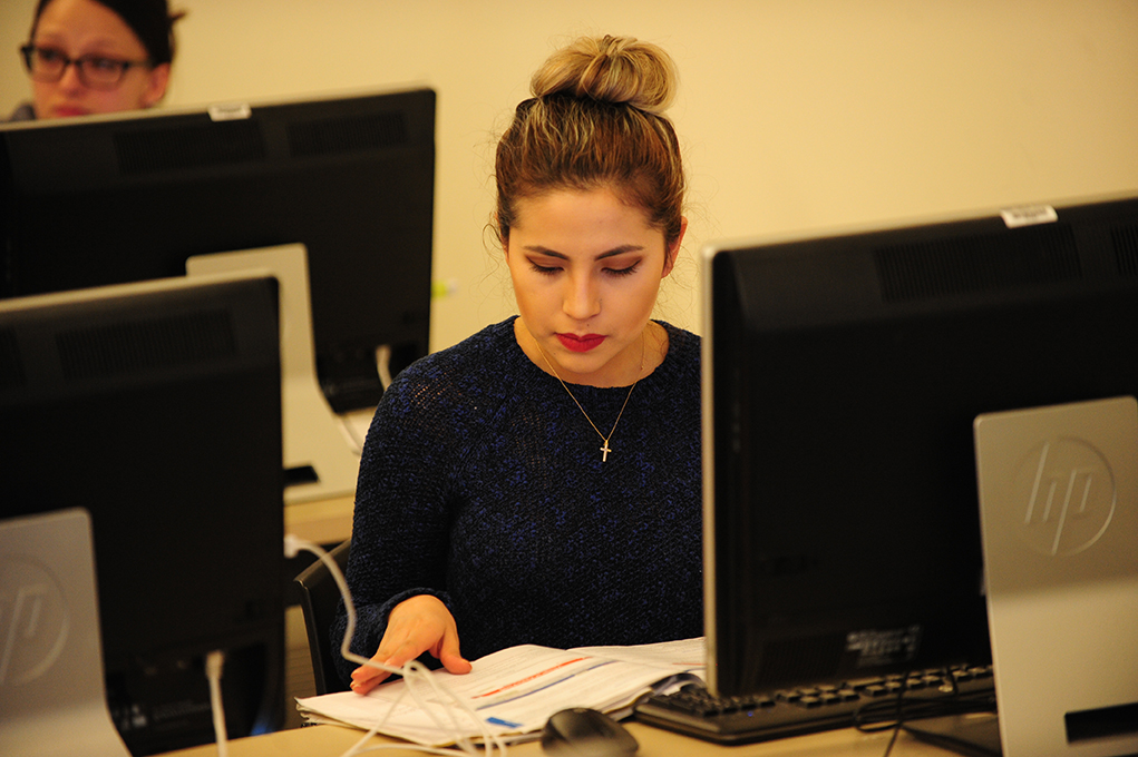 A student reading a textbook in front of a desktop computer.